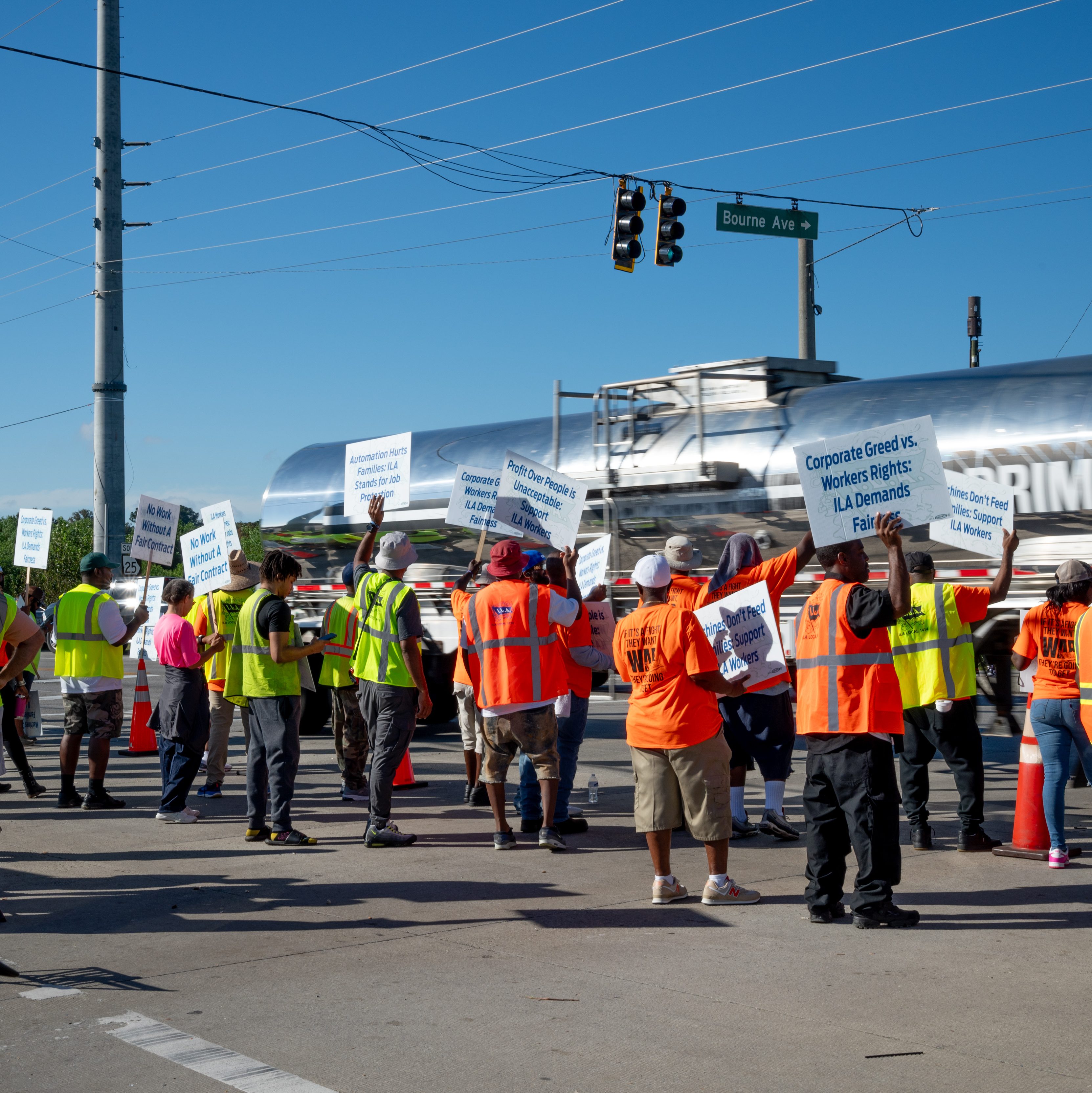 A group of ILA Union workers in safety vests and hats participate in a protest, holding signs that advocate for workers' rights and fair treatment, with a busy road and traffic signal in the background.